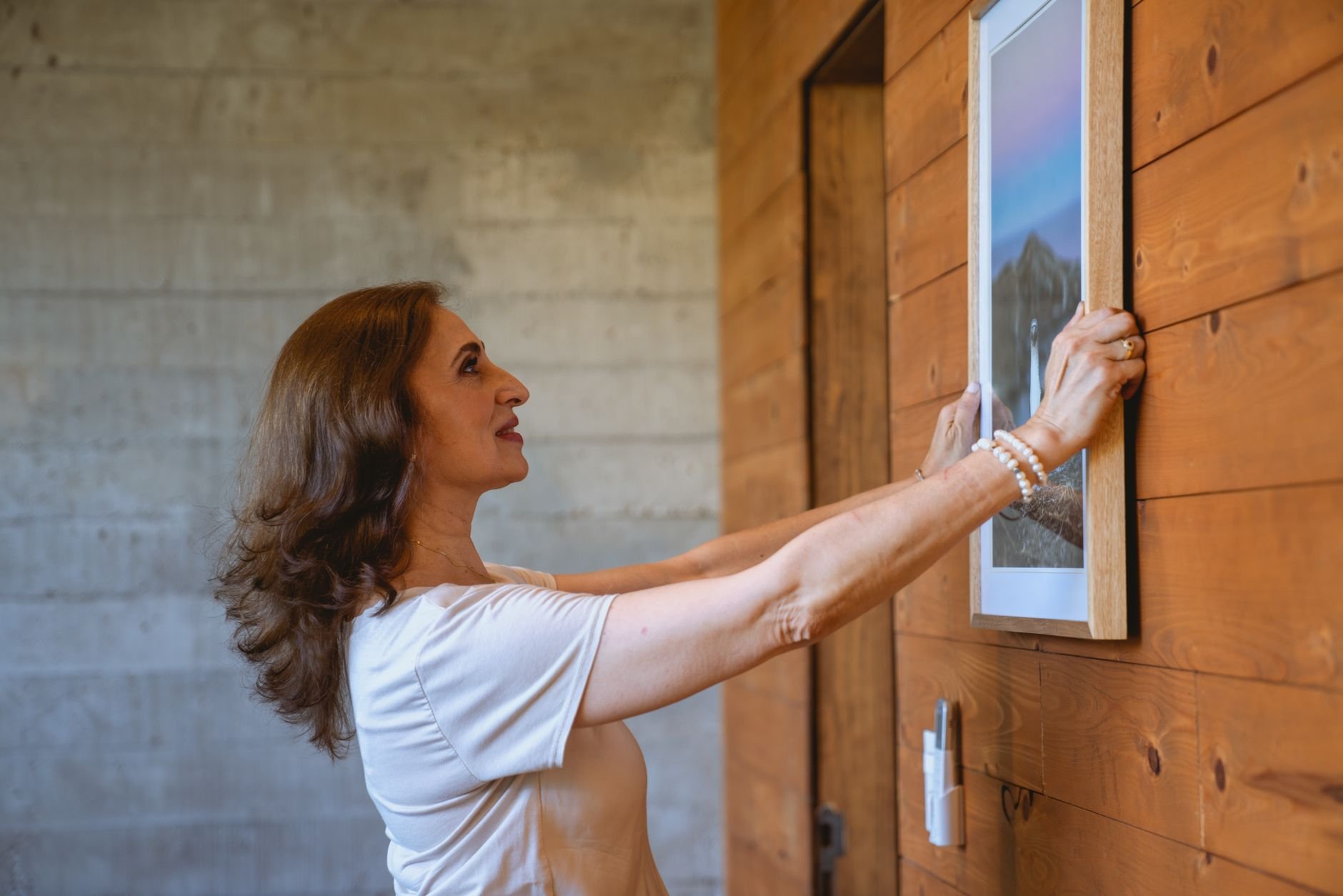 Adult woman decorates home with framed picture on wooden wall, creating a cozy interior.